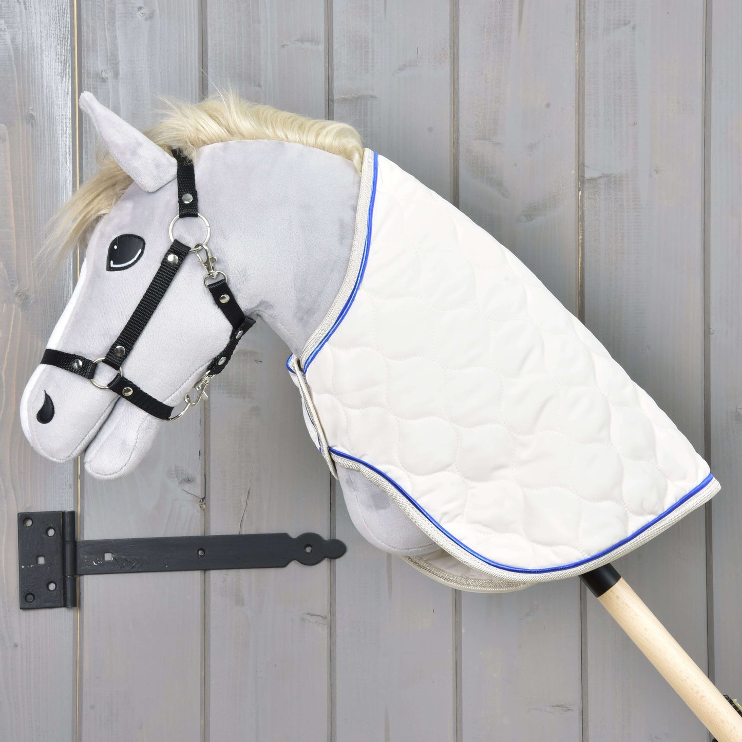 Hobby Horse LarDen se šedou plyšovou hlavou, černou uzdečkou a dekou Cream Pearl s modrým lemováním na dřevěné tyči, zobrazený před šedou dřevěnou stěnou.