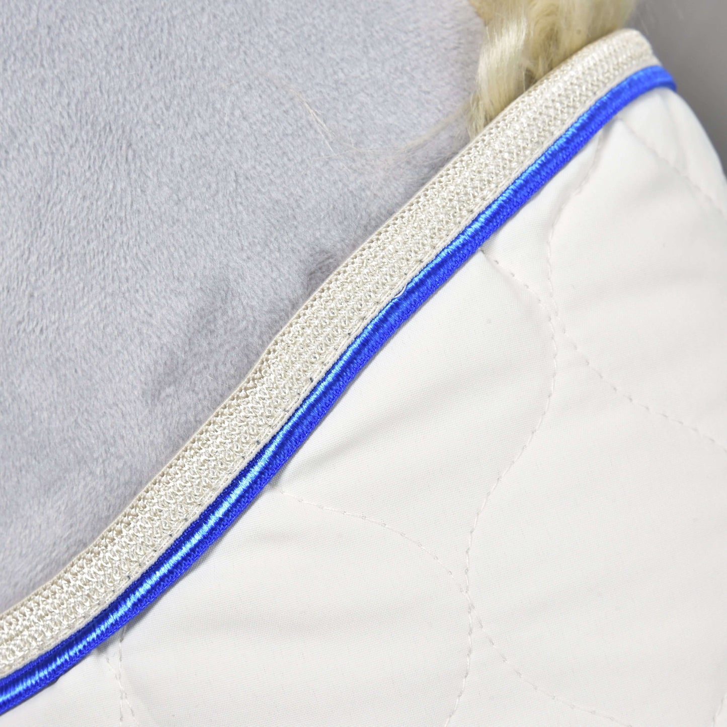 Detail deky Hobby Horse LarDen Cream Pearl s bílou prošívanou látkou, modrou paspulkou a béžovým strukturovaným lemem, položené vedle měkkého světle šedého povrchu.