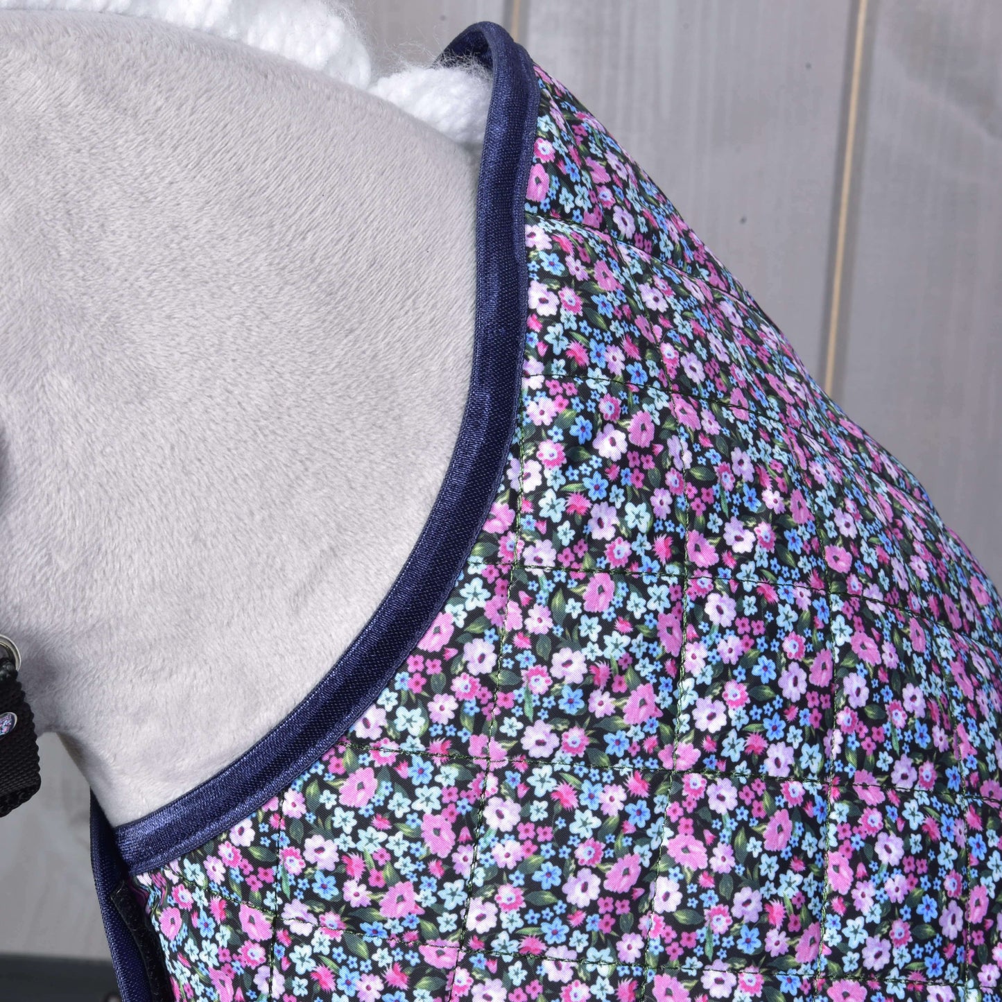 Detail koně s prošívanou dekou Hobby Horse LarDen Blanket Flower s tmavě modrým lemem a malými růžovými, fialovými, bílými a modrými květy na tmavém podkladu. Pozadí tvoří světlé dřevo.