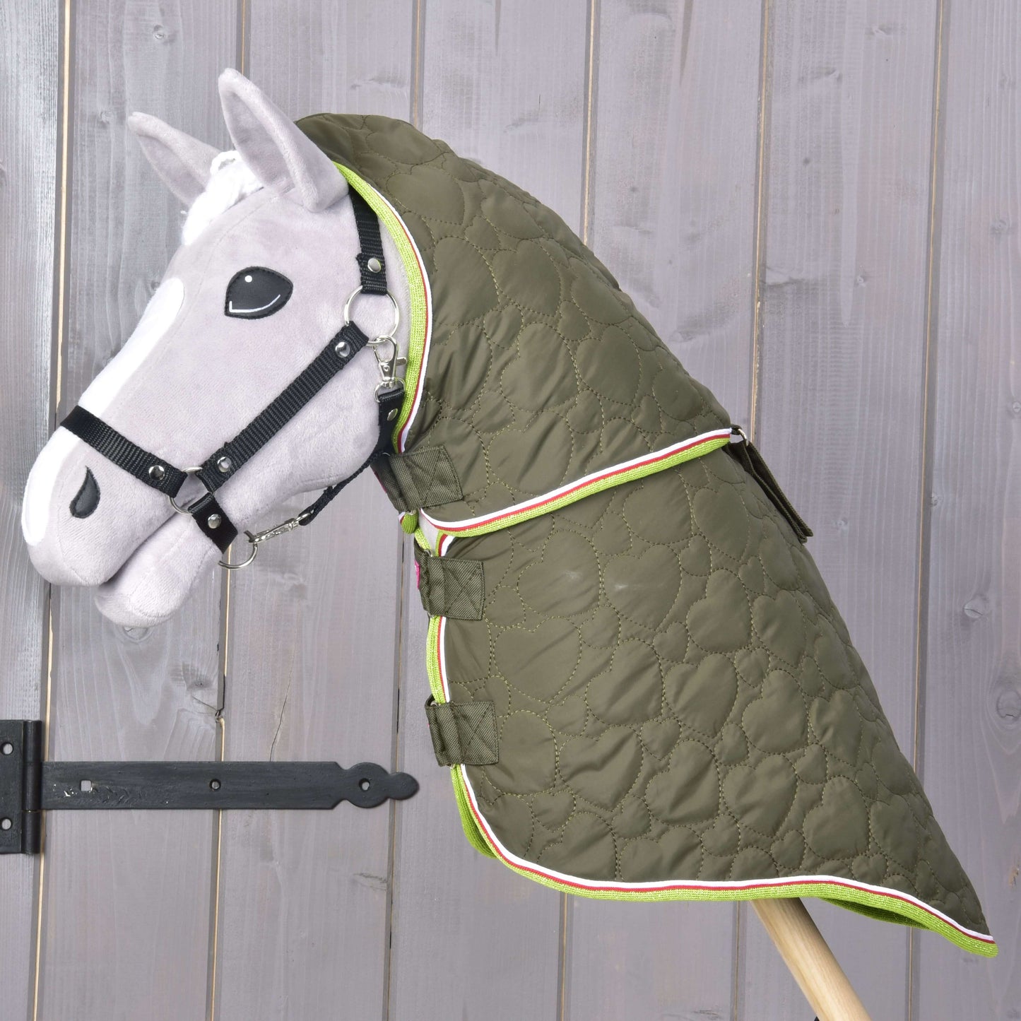 Šedý Hobby Horse s černou uzdečkou má na sobě Deka s odepínatelným krkem Rubby od Hobby Horse LarDen, zelenou kvalitní deku se vzorem srdcí a barevným lemováním, zobrazenou před dřevěnou stěnou.