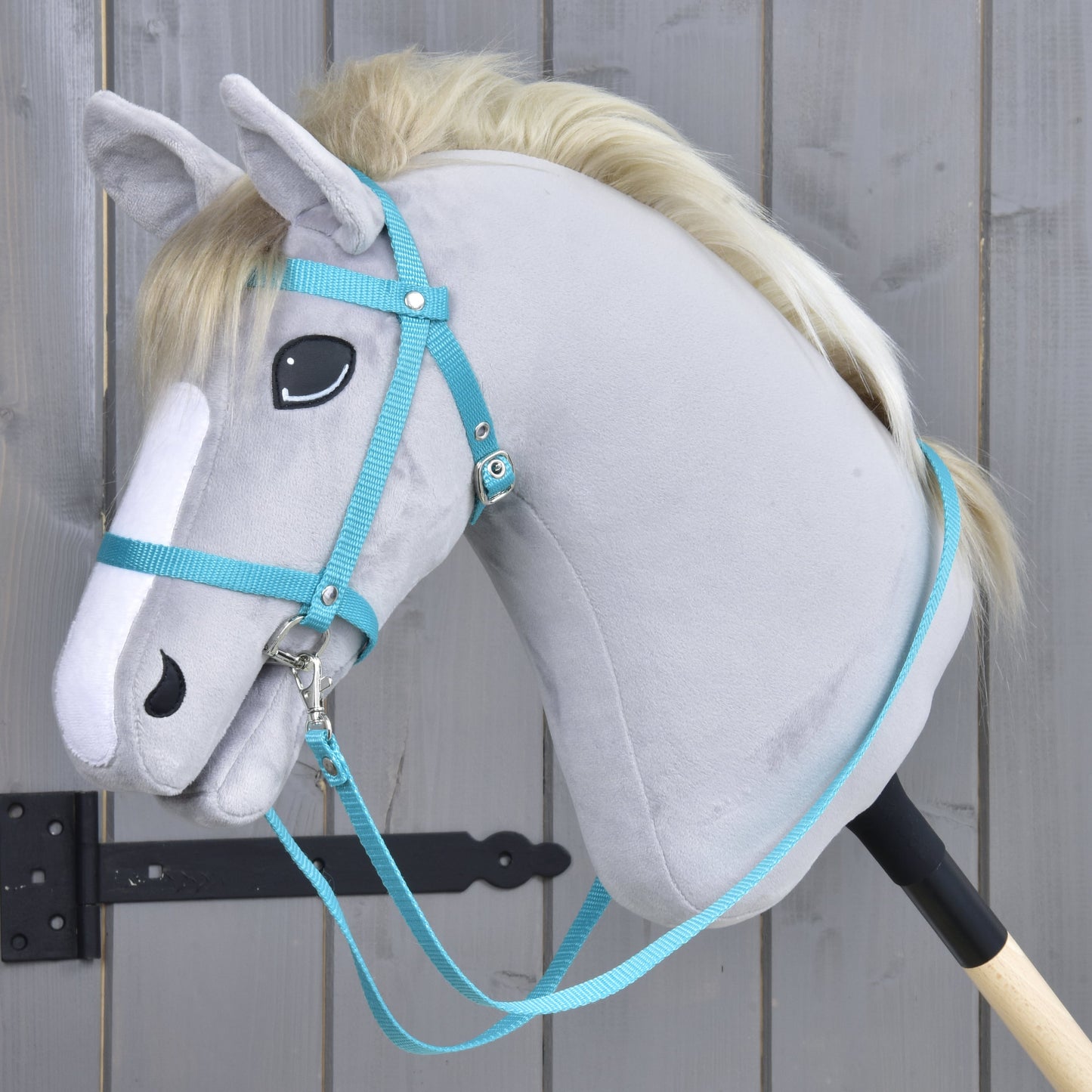 Uzdečka z popruhu Turquoise od Hobby Horse LarDen na šedém plyšovém hobby horse se světlou hřívou, s černou uzdečkou a otěžemi, nasazeném na dřevěné tyči před šedým dřevěným plotem.