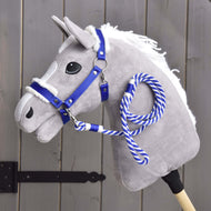 Set Essential Blue od Hobby Horse LarDen se šedým plyšovým Hobby Horse s bílou hřívou, modrou ohlávkou s kožešinovým lemem a modro-bílou provazovou otěží na dřevěné tyči, zobrazený před šedým dřevěným plotem.