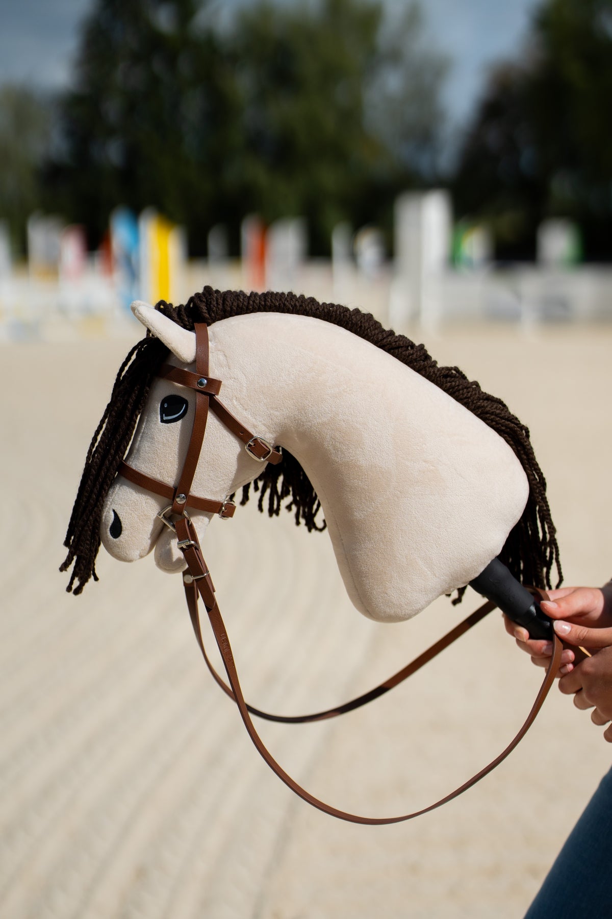 Hlava Hobby Horse Frederik LD s černou ohlávkou od Hobby Horse LarDen držená na pískové jízdárně před rozmazanými barevnými skokovými stojany, krémový plyš, tmavě hnědá přízová hříva a ofina, s kompletní hnědou uzdečkou s nánosníkem