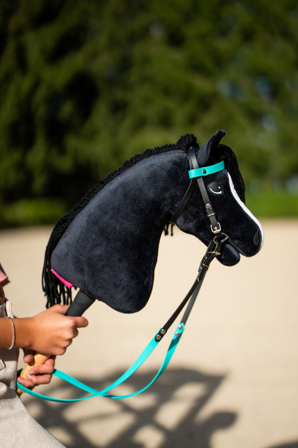 Hlava Hobby Horse Freya s černou ohlávkou od Hobby Horse LarDen držená na pískové jízdárně před tmavými jehličnany, černý plyš, bílá lysina, černá přízová hříva, s kompletní tyrkysovou uzdečkou bez nánosníku