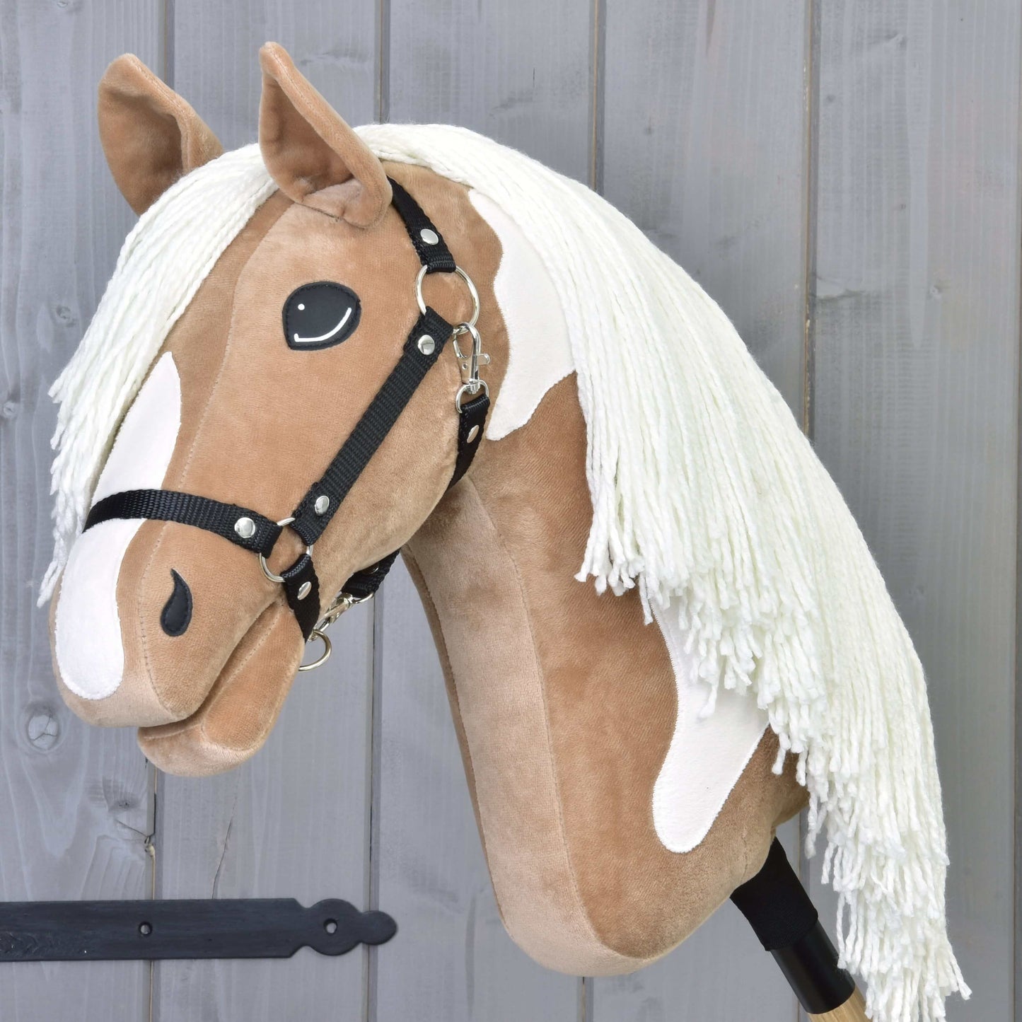 Hobby Horse Fury s černou ohlávkou od Hobby Horse LarDen má plyšový světle hnědý a bílý obličej, dlouhou bílou hřívu, černou ohlávku a je nasazený na tyči, připravený na vyjížďky po stezkách před šedým dřevěným pozadím.