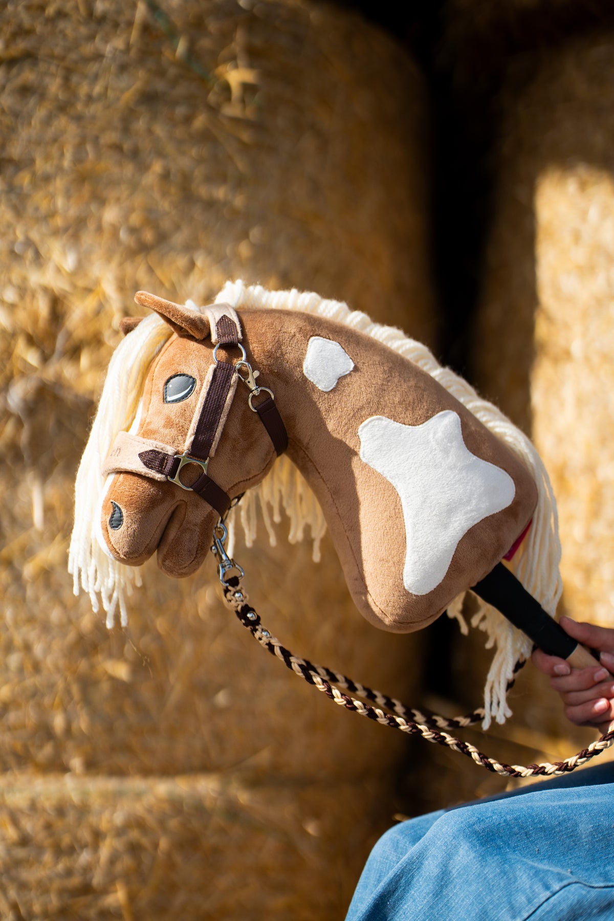 Hlava a krk Hobby Horse Fury s černou ohlávkou od Hobby Horse LarDen před kulatými balíky slámy, plyš palomino s bílými znaky, krémová přízová hříva, s ohlávkou LarDen Brown s kožešinovým lemováním a pleteným vodítkem Bloom Brown