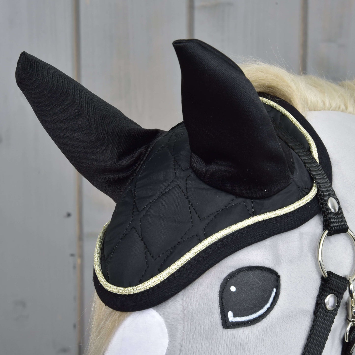 Detail hlavy šedého hračkového koně s černou prošívanou čabrakou Set Majestic od Hobby Horse LarDen se zlatým lemem, vztyčenými ušima a jednoduchou černou uzdečkou, zobrazený na dřevěném pozadí.