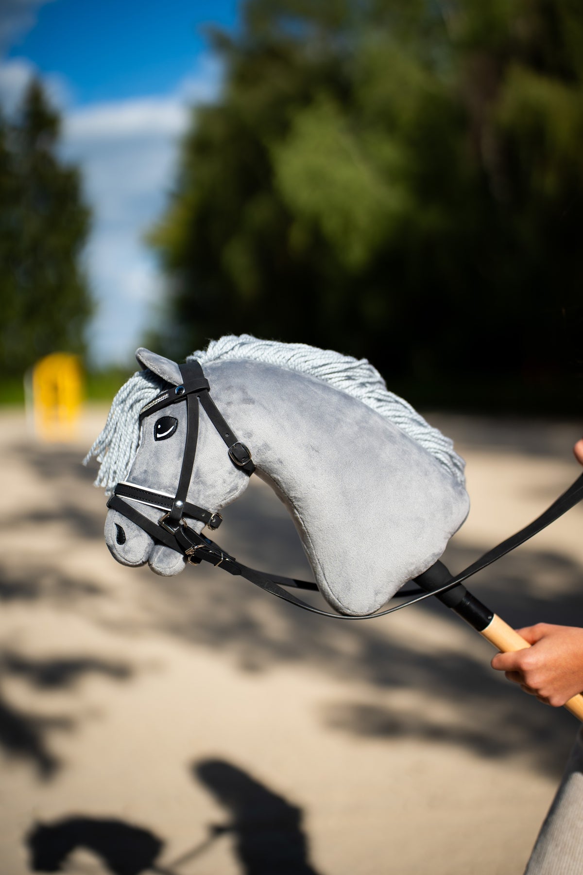 Hlava Hobby Horse Grace s černou ohlávkou od Hobby Horse LarDen držená na pískové jízdárně před tmavými jehličnany, šedý plyš, světle šedá přízová hříva a ofina, s kompletní uzdečkou De Luxe se dvěma čelenkami
