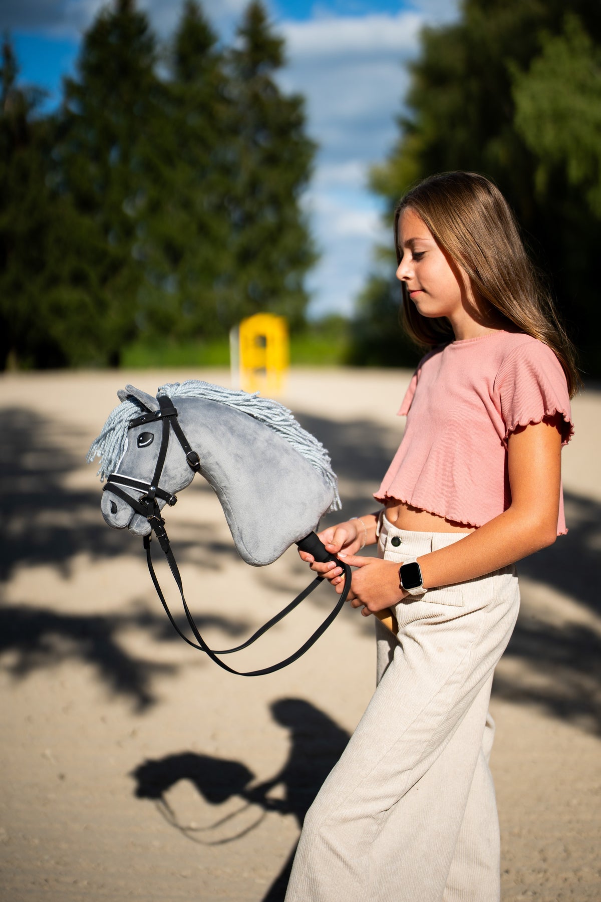 Dívka v růžovém topu vede Hobby Horse Grace s černou ohlávkou od Hobby Horse LarDen přes pískovou jízdárnu, šedá plyšová hlava, světle šedá přízová hříva, s kompletní uzdečkou De Luxe se dvěma čelenkami