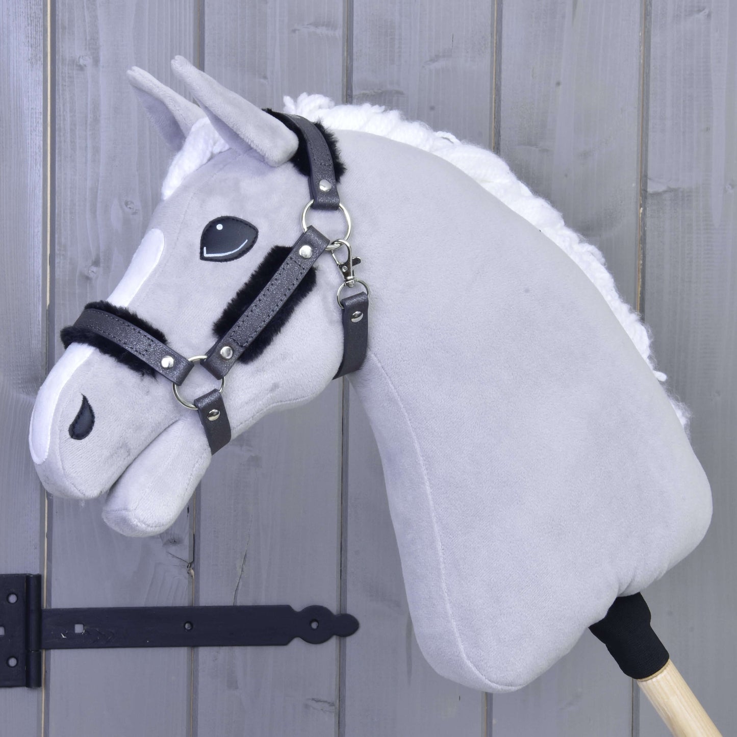 Ohlávka s beránkem z koženky Graphite Full od Hobby Horse LarDen na dřevěné tyči, opřená o světle šedou dřevěnou stěnu, se šedou plyšovou hlavou s bílou hřívou, černou uzdečkou a ohlávkou z koženky.