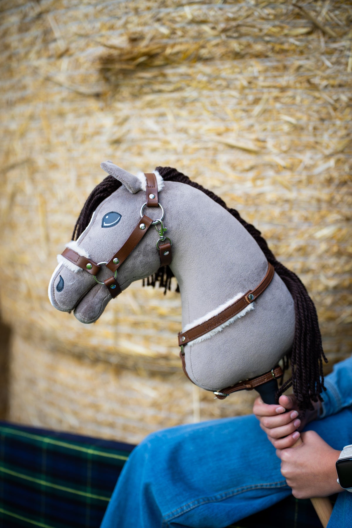 Hlava Hobby Horse Guccini od Hobby Horse LarDen před balíky slámy, z šedého plyše s bílým pruhem na nose a tmavě hnědou přízovou hřívou, s Ohlávkou Walnut s kožešinovým podšitím a náprsníkem Walnut s kožešinou