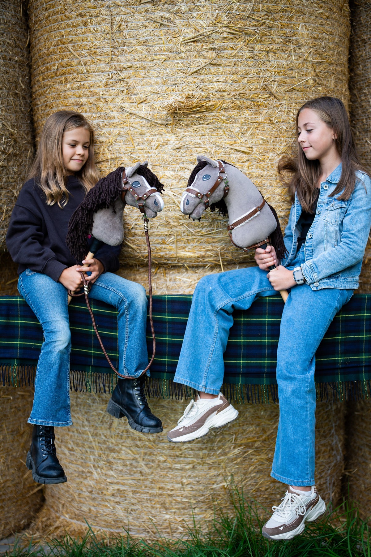 Dvě dívky sedí na balících slámy na tartanové dece a každá drží Hobby Horse Guccini od Hobby Horse LarDen, šedou hlavu ve stylu holštýnského koně s bílým pruhem na nose a dlouhou tmavě hnědou přízovou hřívou, s Ohlávkou Walnut s kožešinovým podšitím, náprsníkem Walnut s kožešinou a Vodítkem Vivaldi