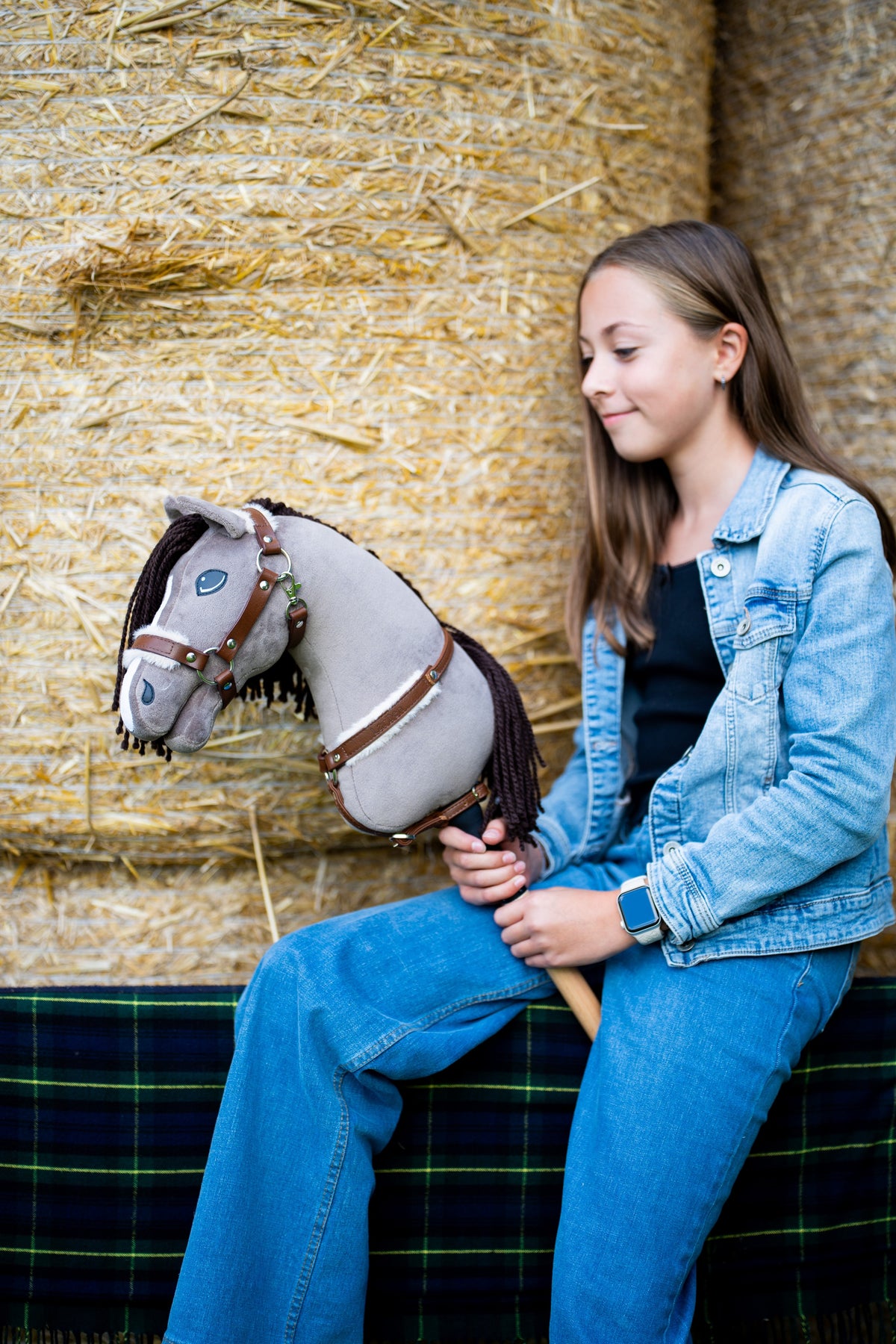 Dívka v džínové bundě sedí na balících slámy a drží Hobby Horse Guccini od Hobby Horse LarDen, šedou hlavu s bílým pruhem na nose, dlouhou tmavě hnědou přízovou hřívou a dřevěnou tyčí, s Ohlávkou Walnut s kožešinovým podšitím a náprsníkem Walnut s kožešinou