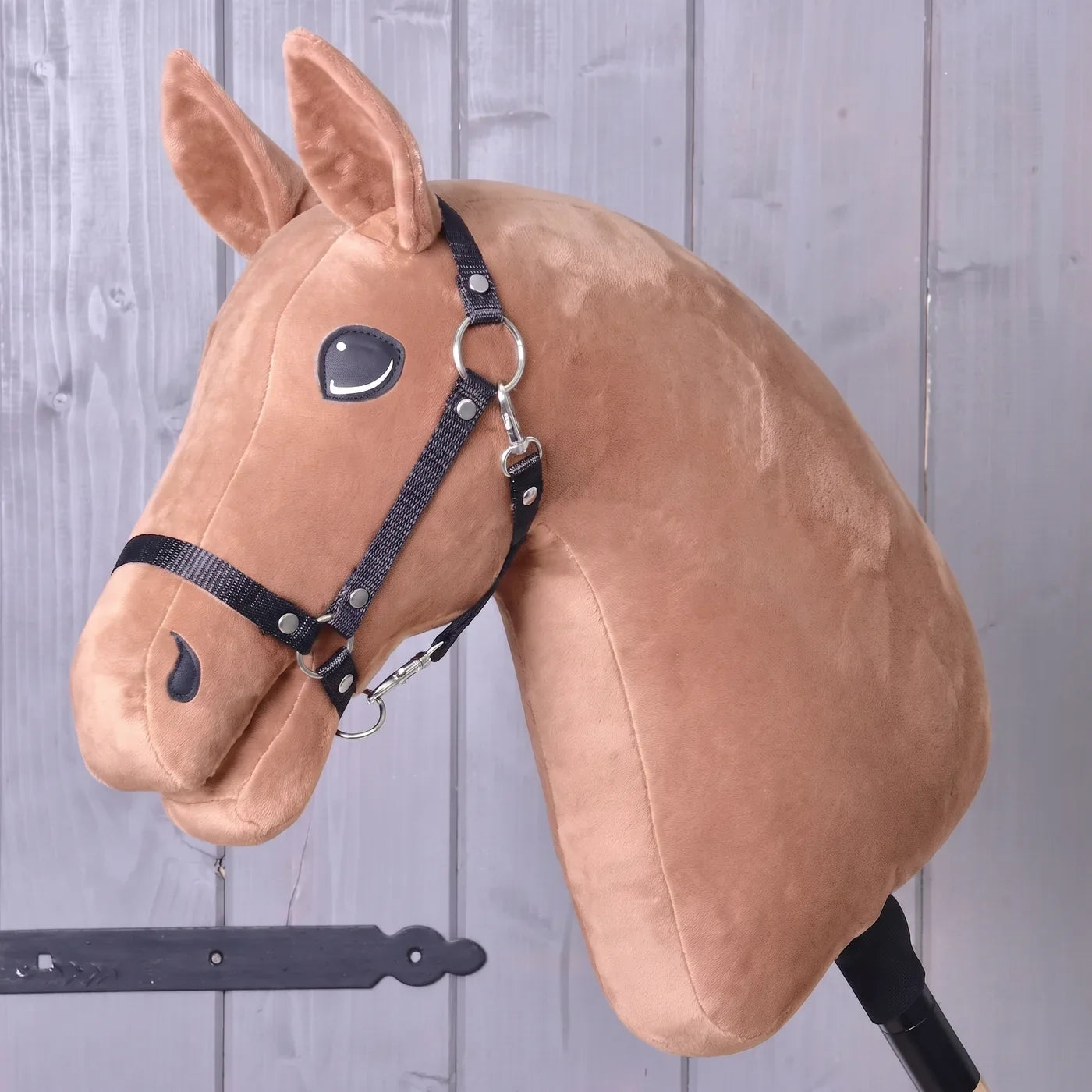 Sestav si svého koně – plyšová hlava Hobby Horse s uzdečkou, nasazená na tyči, opřená o dřevěnou stěnu; ideální inspirace pro vytvoření vlastního jedinečného koně pomocí konfigurátoru „Build your horse“ od Hobby Horse LarDen před dokončením nákupu.