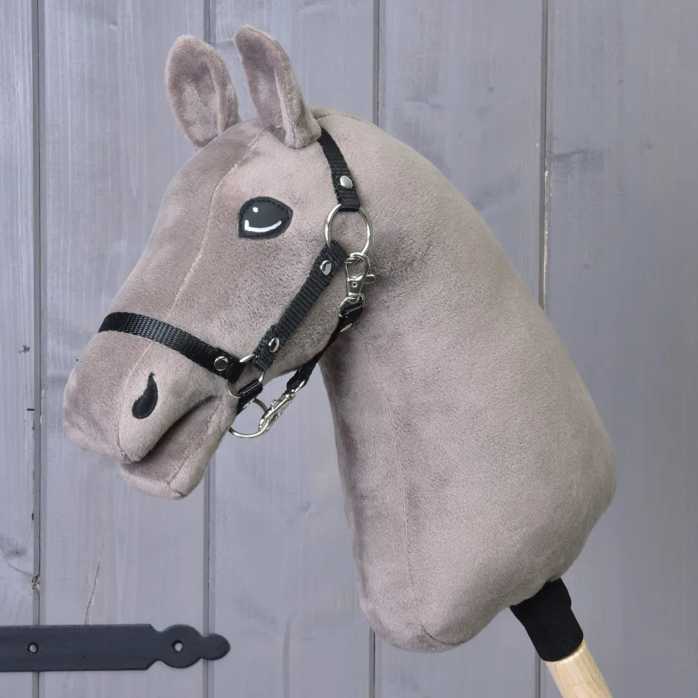Build your horse, plyšová hlava Hobby Horse v taupe barvě s černou přízovou hřívou a uzdečkou na dřevěné tyči, zobrazená před šedou dřevěnou stěnou.