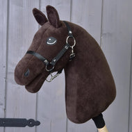 Plyšová tmavě hnědá hlava Hobby Horse Build your horse s černým postrojem a uzdečkou, upevněná na dřevěné tyči, zobrazená před šedými dřevěnými prkny.