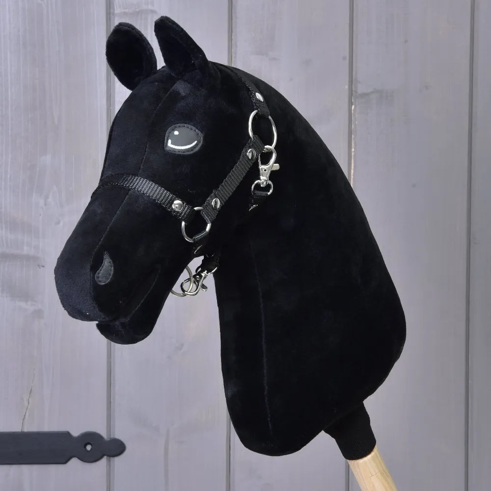 Build your horse, černá plyšová vlastní hlava Hobby Horse s tříbarevnou hřívou z umělé kožešiny, uzdečkou a otěžemi na dřevěné tyči, na pozadí šedého dřevěného panelu – přizpůsobte si ji přesně podle svých představ!