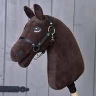 Build your horse, plyšový tmavě hnědý Hobby Horse s černou uzdečkou, otěžemi a přízovou hřívou na tyči, zobrazený na pozadí šedého dřevěného obložení.