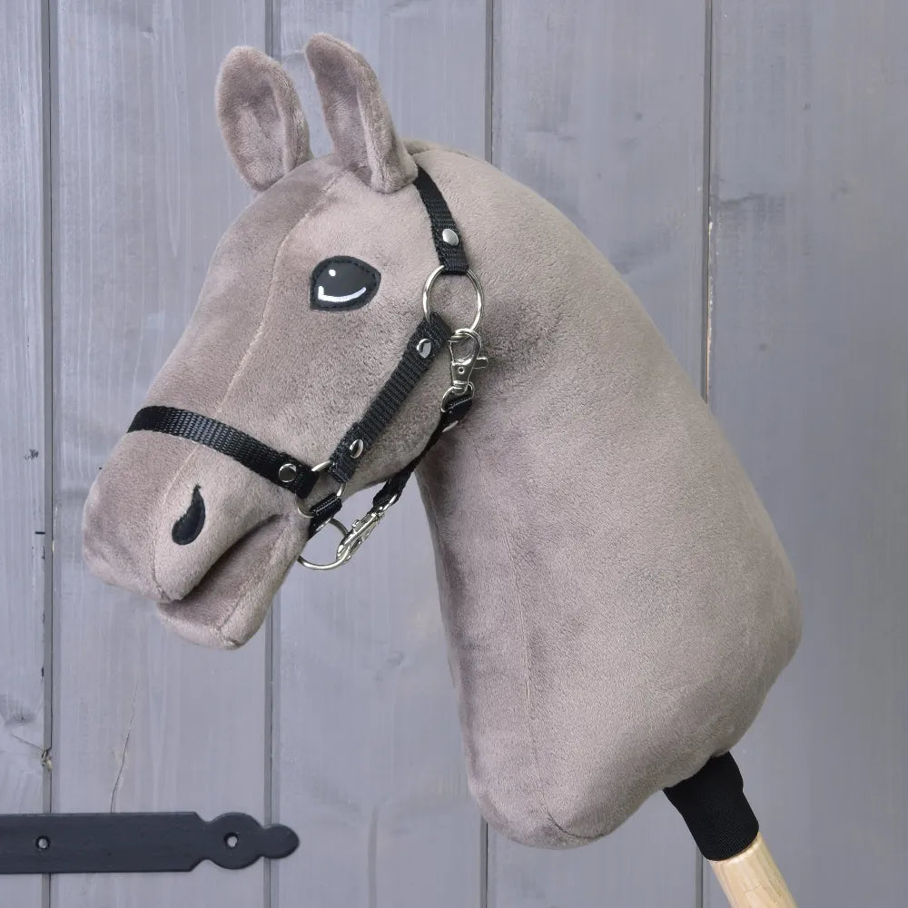 Build your horse od related_to_12441055625556, plyšový taupe Hobby Horse s hnědo-modrýma očima, černou uzdečkou a otěžemi, upevněný na dřevěné tyči, zobrazený na šedém dřevěném pozadí.