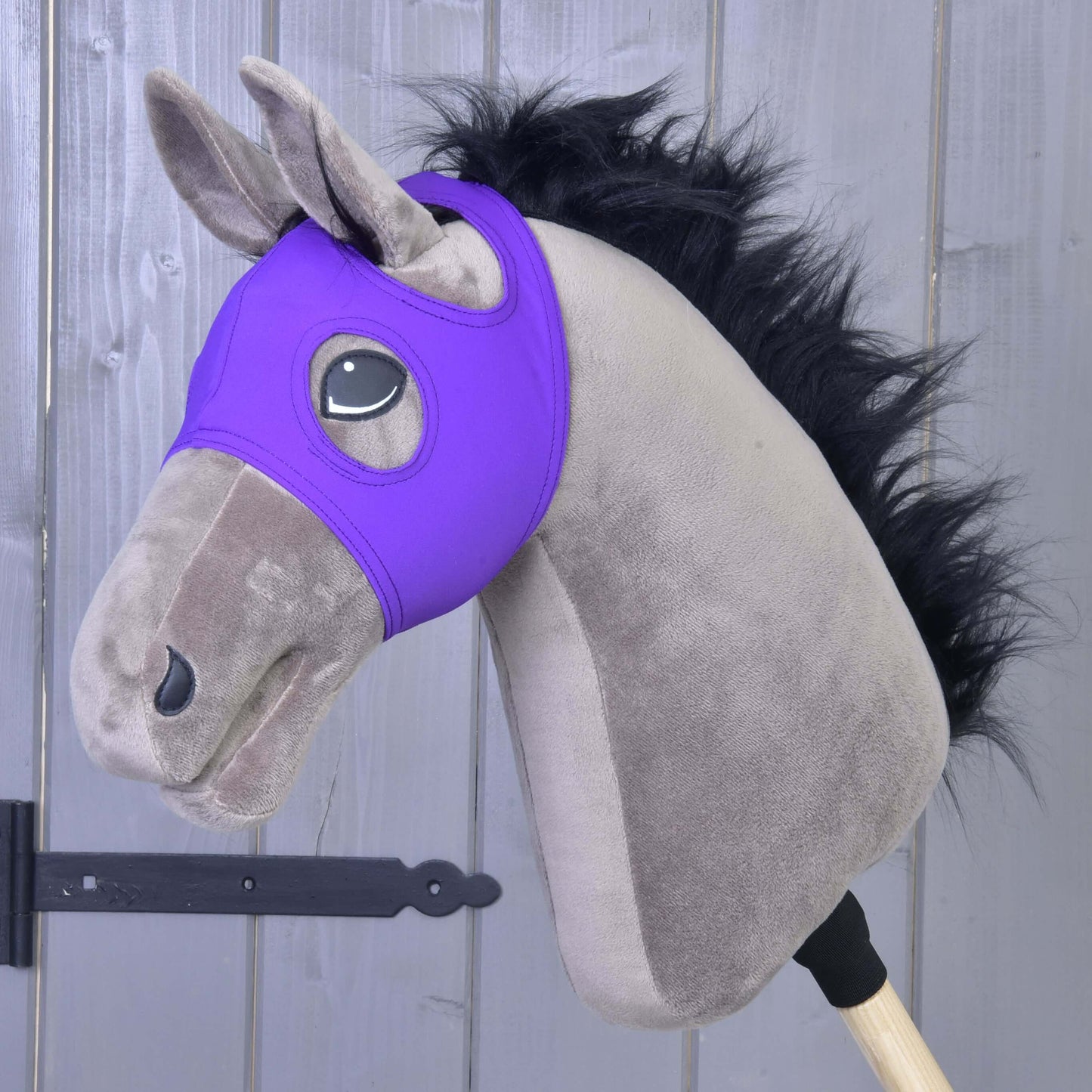 Plyšový Hobby Horse s černou hřívou, šedou hlavou a Maskou bez uší Indigo od Hobby Horse LarDen v indigové barvě, vyrobenou z pratelného materiálu PES, nasazený na dřevěné tyči na pozadí šedé dřevěné stěny.