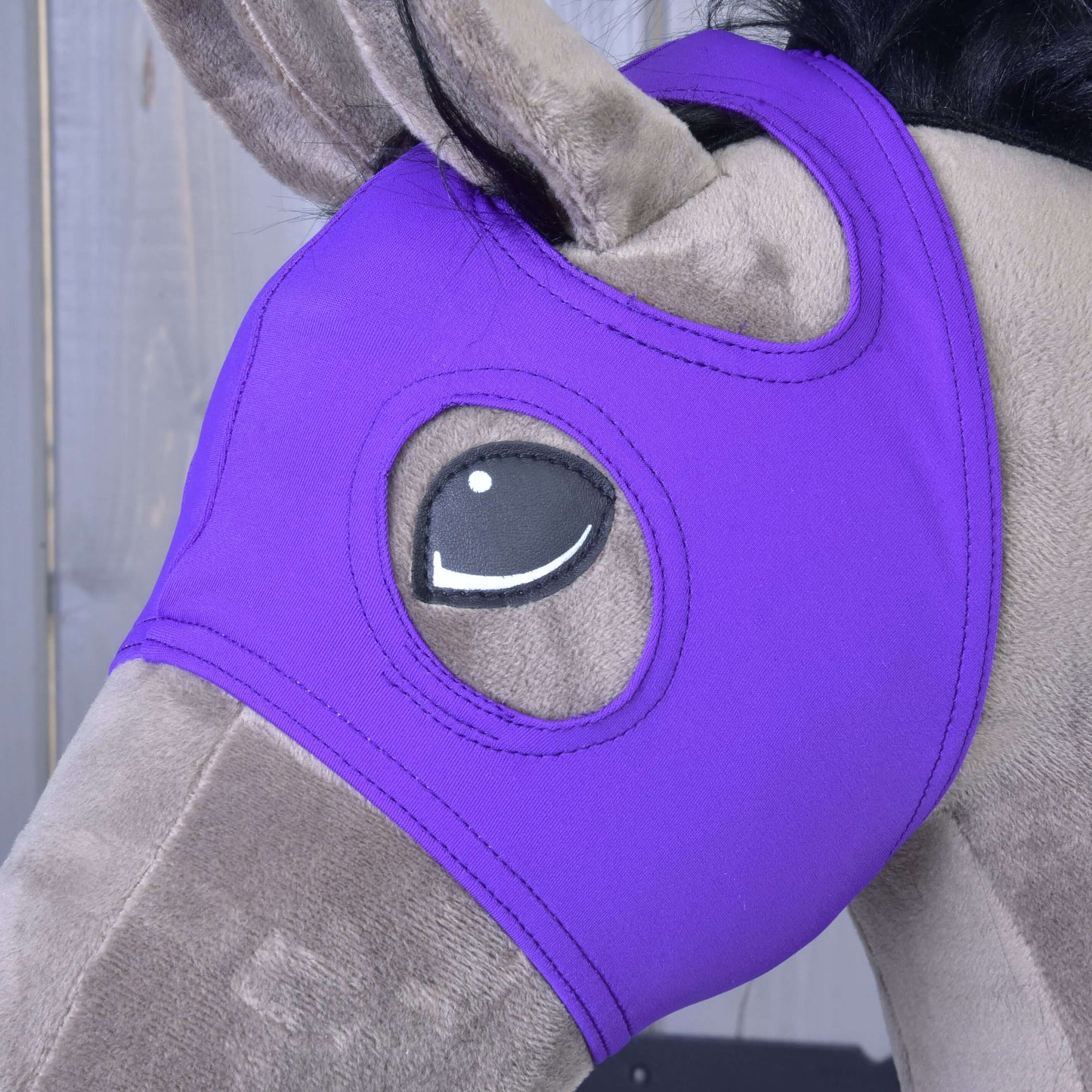 Plyšový hračkový kůň má nasazenou Masku bez uší Indigo od Hobby Horse LarDen, vyrobenou z pratelného PES, zakrývající hlavu s otvory pro oči, na dřevěném pozadí.