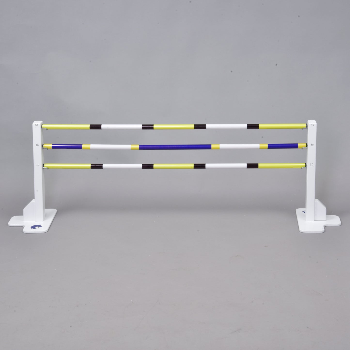 Bílý kůň skáče přes set překážky kolmák 50 cm + 2 ks bariérky od Hobby Horse LarDen s extra žlutými, bílými, modrými a černými tyčemi na šedém pozadí. Ideální pro aktivity Hobby Horsing uvnitř i venku.