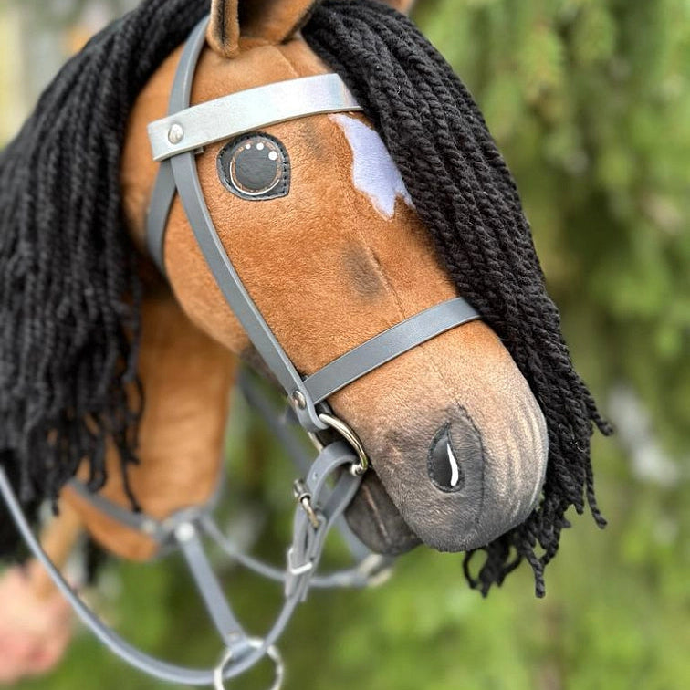 Detail ručně vyrobeného Hobby Horse s černou přízovou hřívou, hnědým tělem, bílou skvrnou nad jedním okem a šedou uzdečkou na rozmazaném zeleném venkovním pozadí – dokonalý doplněk pro dobrodružství s Hobby Horsing.