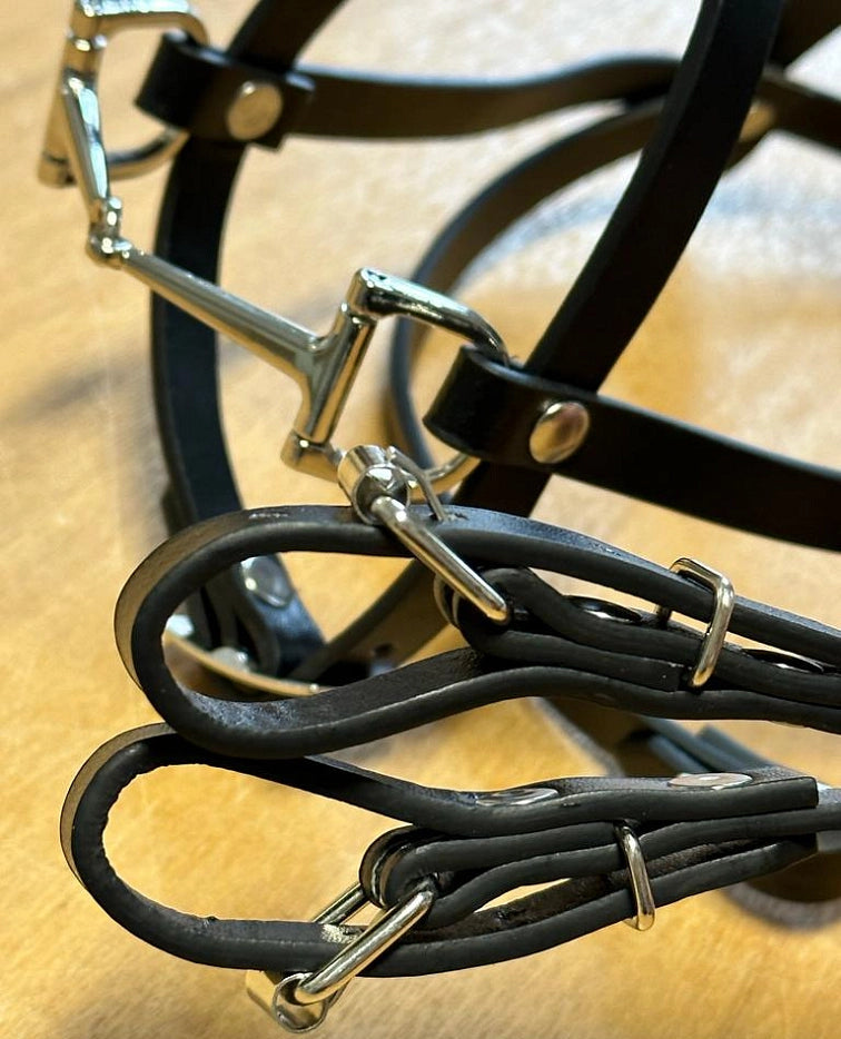 Detail černé kožené koňské uzdečky a otěží s lesklými stříbrnými kovovými přezkami a kroužky, ideální pro výbavu na Hobby Horsing, položených na světle hnědém povrchu.