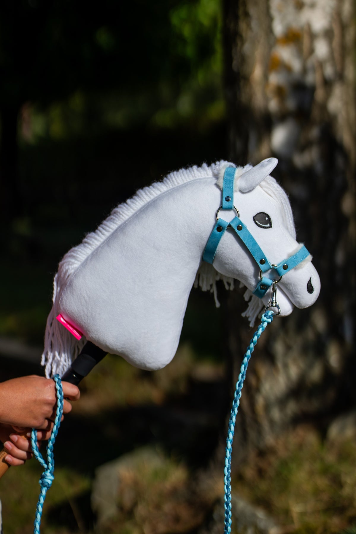 Hlava Hobby Horse Lady s černou ohlávkou od LarDen je zvednutá v kropenatém stínu, s bílým plyšem, tmavým okem a bílou přízovou hřívou, s koženkovou kožešinovou ohlávkou Pastell Blue a pleteným vodítkem Double Blue