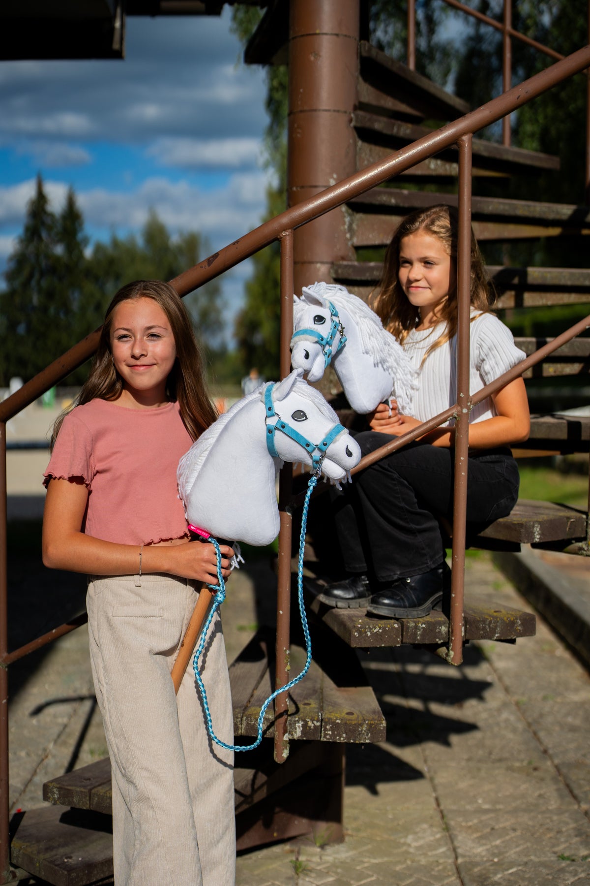 Dvě dívky s bílými Hobby Horses od LarDen stojí na rezavém kovovém schodišti, vpředu je Hobby Horse Lady s černou ohlávkou, s bílou plyšovou hlavou a bílou přízovou hřívou, s koženkovou kožešinovou ohlávkou Pastell Blue a pleteným vodítkem Double Blue