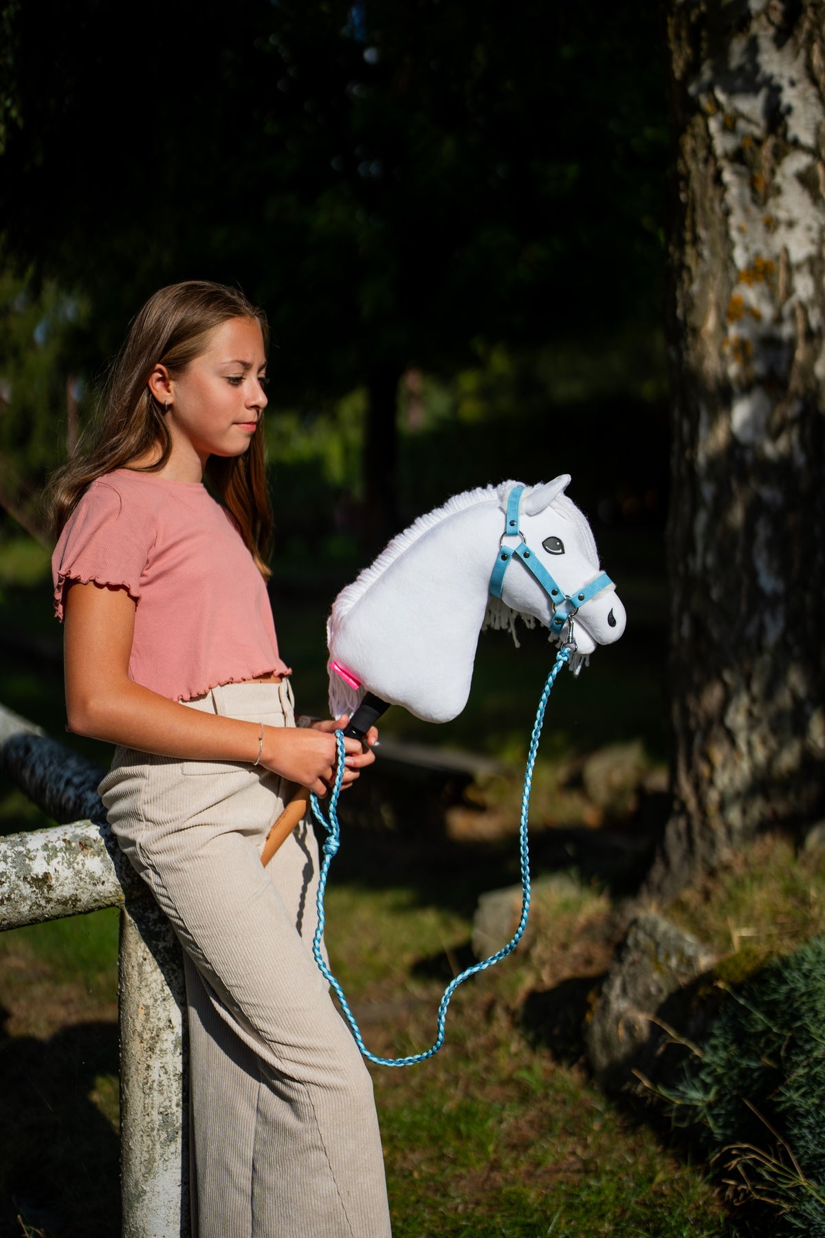 Dívka v růžovém topu stojí u mechem porostlého stromu a drží Hobby Horse Lady s černou ohlávkou od Hobby Horse LarDen, vodítko visí ve smyčce, bílá plyšová hlava, bílá přízová hříva, s ohlávkou Pastell Blue s kožešinou z koženky a pleteným vodítkem Double Blue