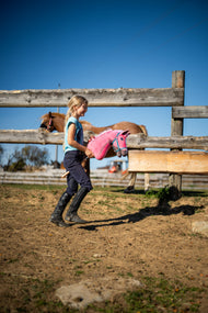 Dívka v jezdeckém oblečení prochází písčitým výběhem s Hobby Horse LarDen Pinkie s třpytivou ohlávkou od Hobby Horse LarDen s růžovou plyšovou hlavou a růžovobílou hřívou z příze, s tyrkysovou Ohlávkou
