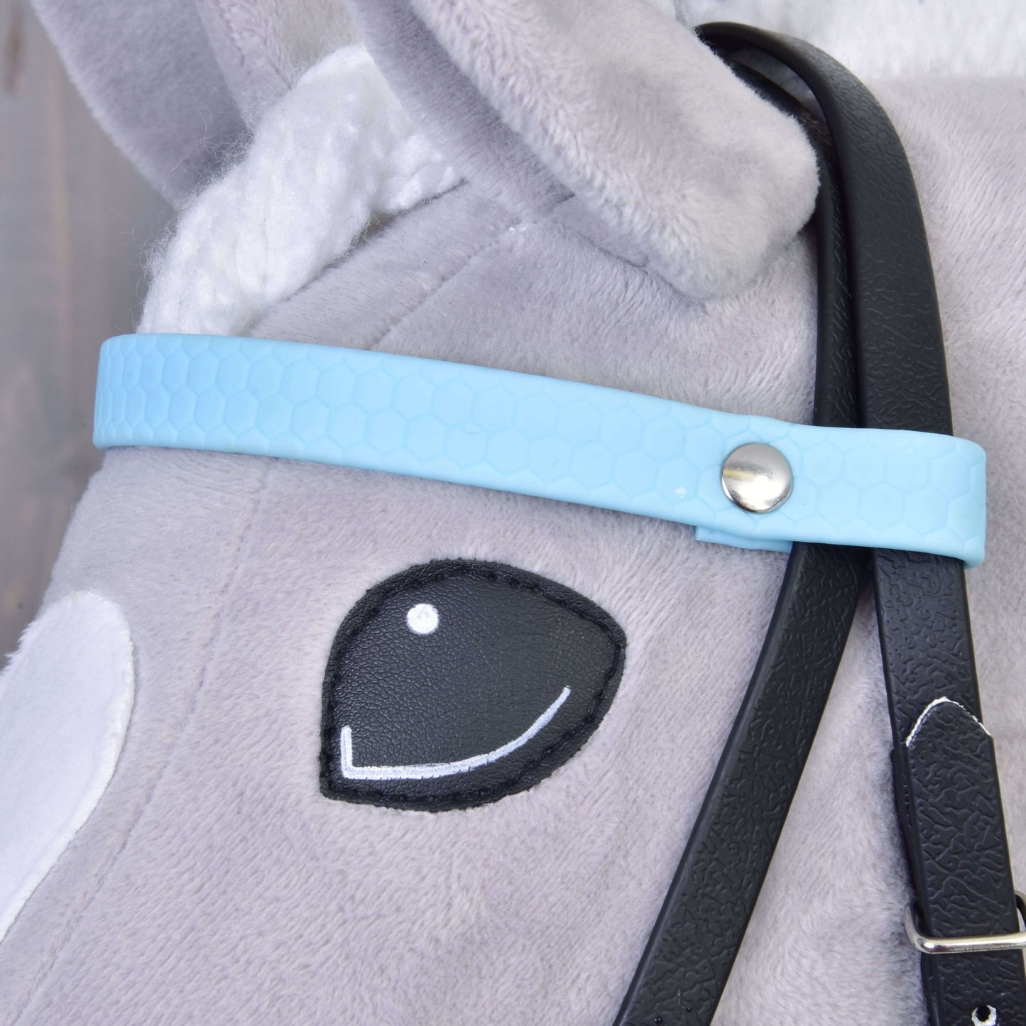 Detail obličeje šedého plyšového hračkového koně s vyšitým černým okem, bílým uchem a Kompletní uzdečkou bez nánosníku Light Blue od Hobby Horse LarDen, která má světle modrý řemínek se stříbrným drukem a odnímatelné černé otěže.