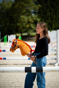 Dívka v černém topu stojí za skokovou tyčí na pískové jízdárně a drží Hobby Horse Little Angel s černou ohlávkou od Hobby Horse LarDen, ryzá plyšová hlava, jasně červená přízová hříva, s ohlávkou Espresso s kožešinou z koženky