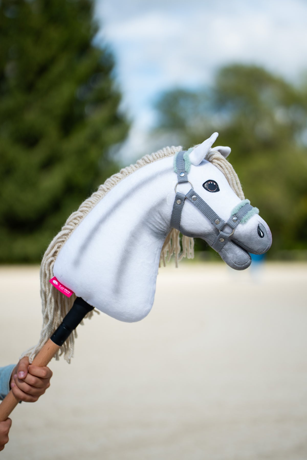 Hlava Hobby Horse Little Lady s černou ohlávkou od LarDen je zvednutá na pískové jízdárně, za ní jsou jehličnany, s bílým plyšem, krémovou přízovou hřívou a ofinou, se šedou ohlávkou s mátovým kožíškem