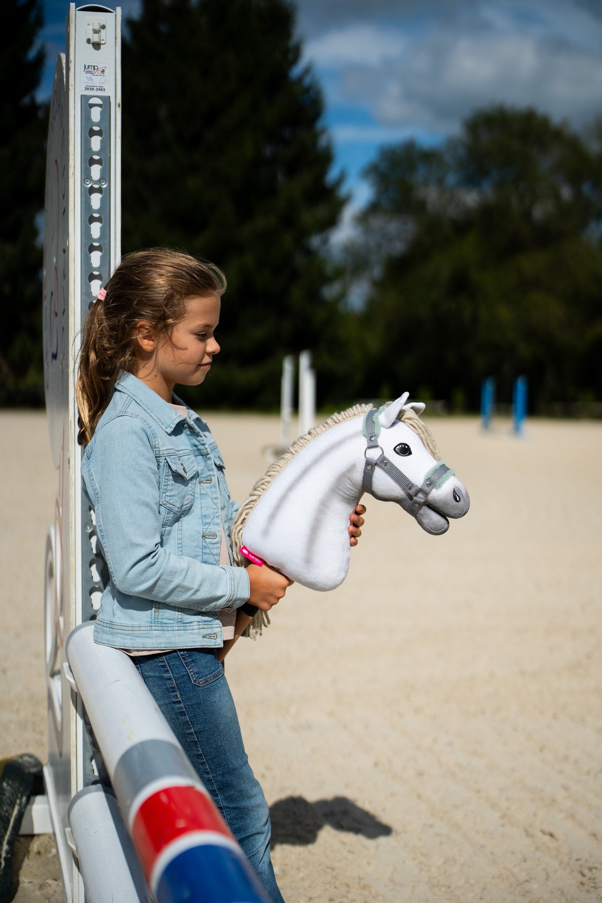 Dívka v džínové bundě stojí u skokového stojanu na pískové jízdárně a drží Hobby Horse Little Lady s černou ohlávkou od Hobby Horse LarDen, bílá plyšová hlava, krémová přízová hříva, se šedou ohlávkou s mátovou kožešinou