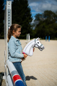 Dívka v džínové bundě stojí u skokového stojanu na pískové jízdárně a drží Hobby Horse Little Lady s černou ohlávkou od Hobby Horse LarDen, bílá plyšová hlava, krémová přízová hříva, se šedou ohlávkou s mátovou kožešinou