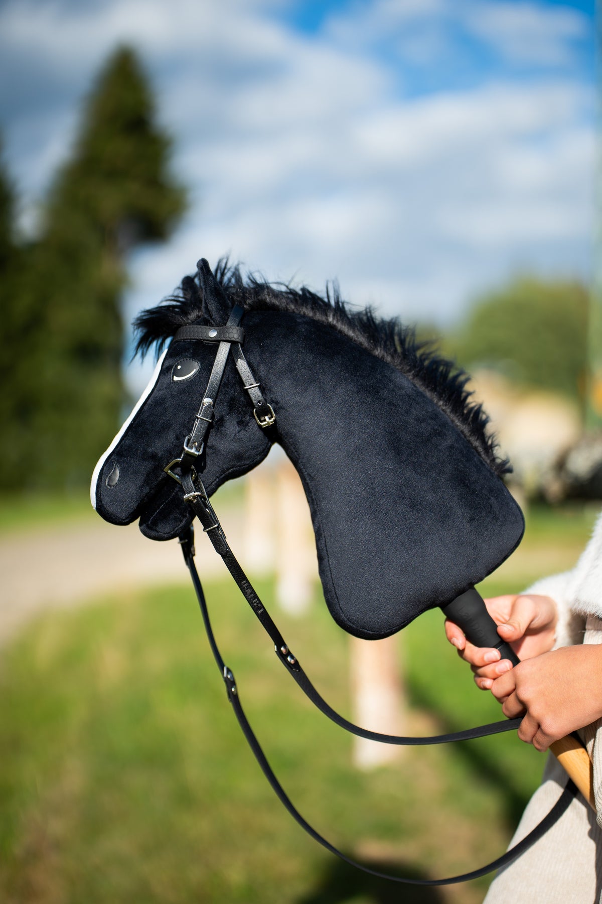 Hlava a krk Hobby Horse Loki s černou ohlávkou od LarDen jsou zvednuté na louce pod modrou oblohou, s černým plyšem, bílou lysinou a černou přízovou hřívou, s kompletní uzdečkou Pure Black bez nánosníku