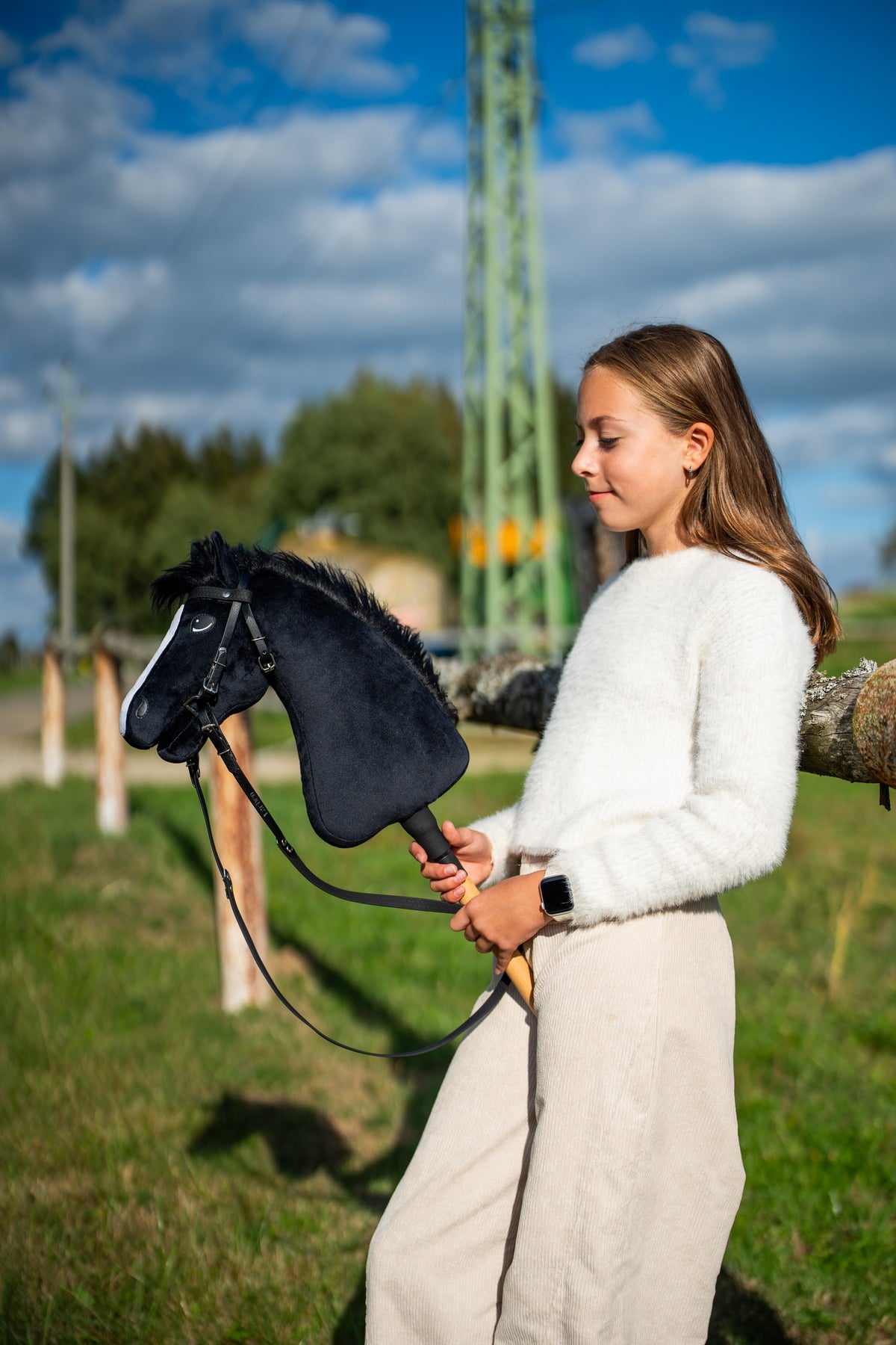 Dívka v bílém svetru stojí na slunné louce a drží Hobby Horse Loki s černou ohlávkou od LarDen, s černou plyšovou hlavou, bílou lysinou a černou přízovou hřívou, s kompletní uzdečkou Pure Black bez nánosníku