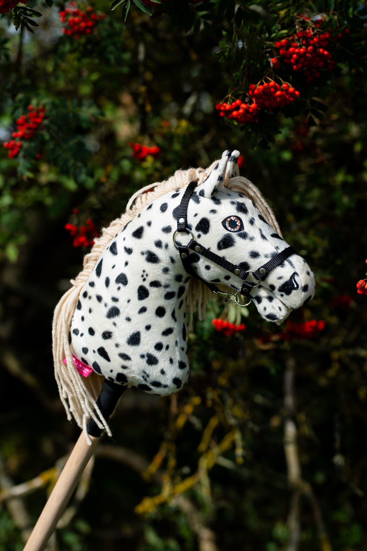 Hobby Horse Lord Valentine s černou ohlávkou od LarDen stojí před jeřábem s trsy červených bobulí, s bílou plyšovou hlavou s černými leopardími skvrnami, krémovou přízovou hřívou a černou ohlávkou se stříbrným kováním
