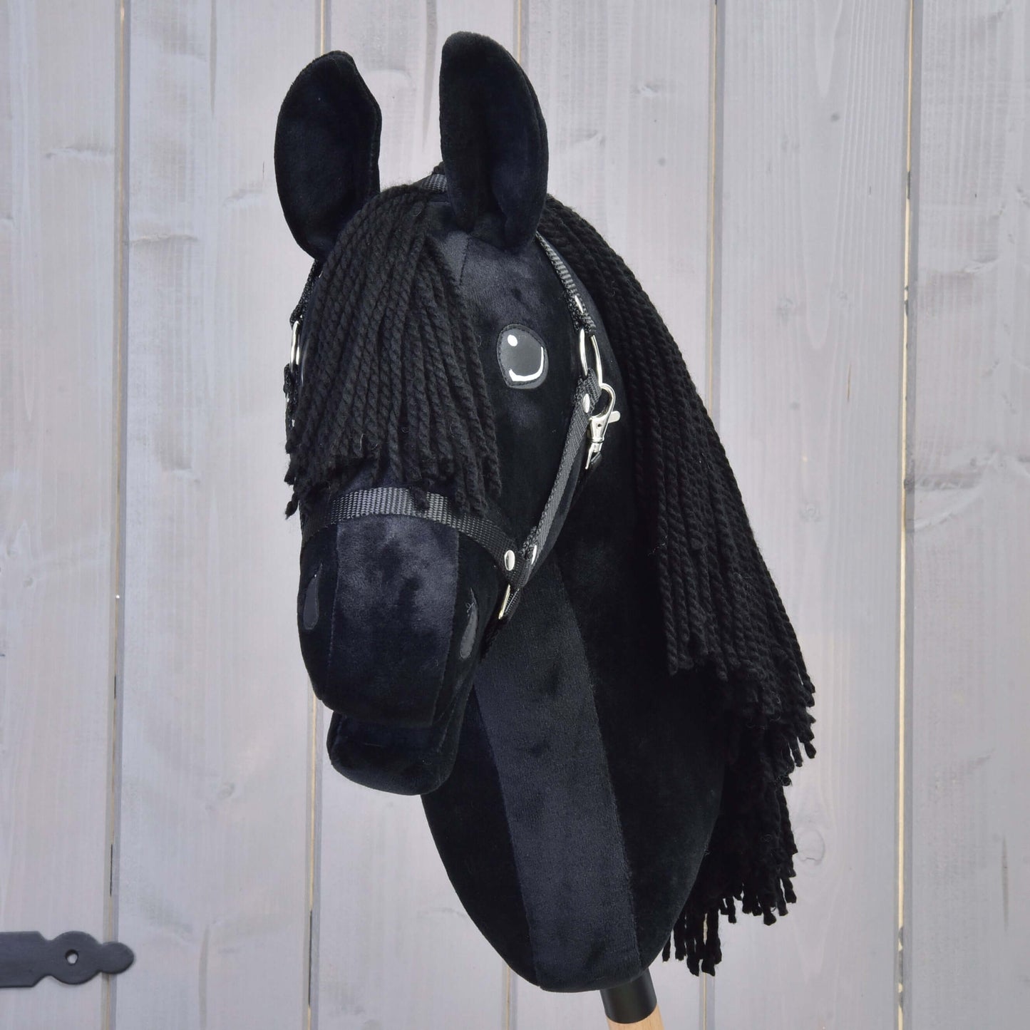 Hobby Horse Mahal s černou ohlávkou od Hobby Horse LarDen má černou plyšovou hlavu inspirovanou fríským koněm s přízovou hřívou a uzdečkou, upevněnou na tyči před světlým dřevěným pozadím.