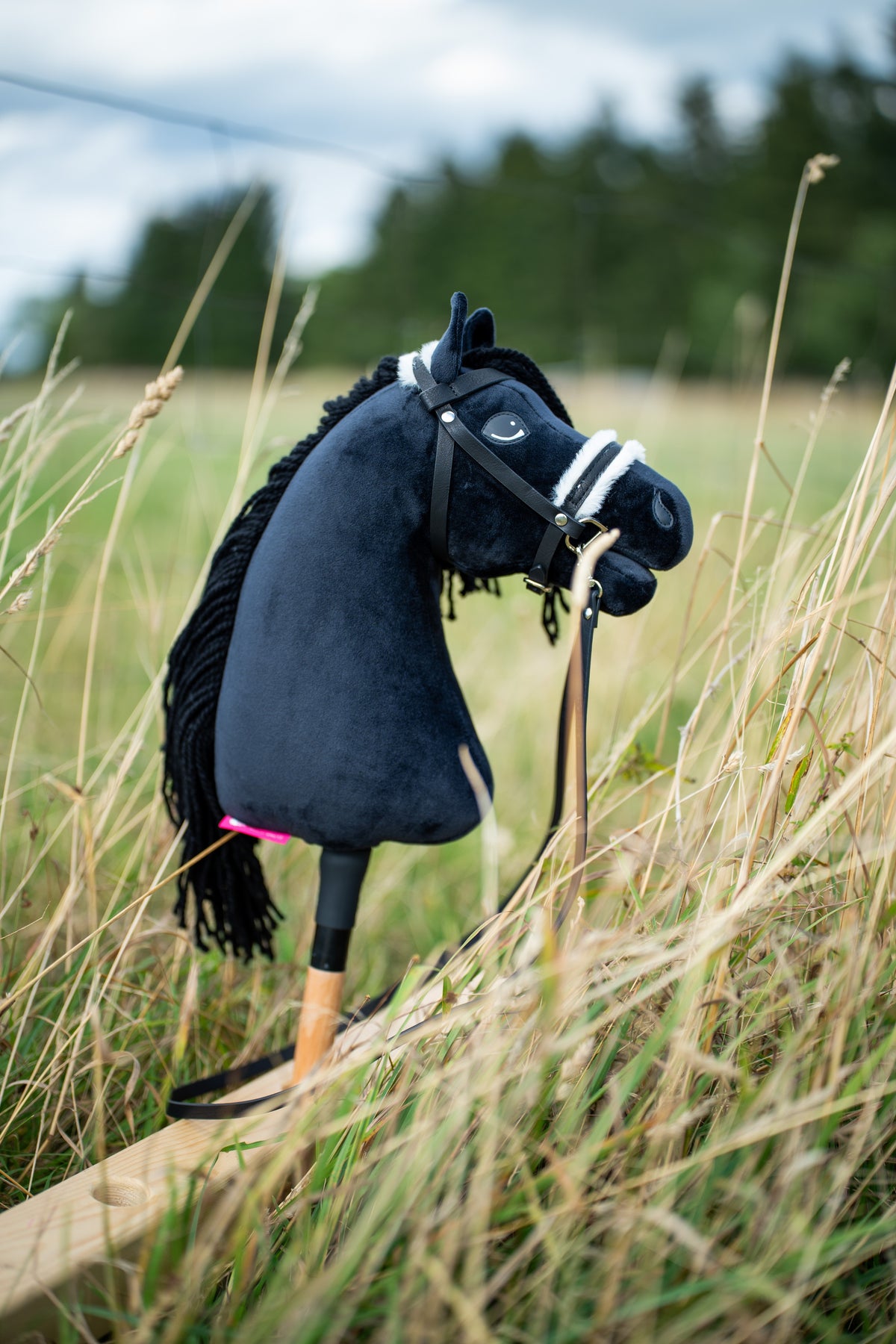 Hobby Horse Mahal s černou ohlávkou od LarDen stojí ve vysoké suché trávě na louce, s černou plyšovou hlavou, černou přízovou hřívou a dřevěnou tyčí, s kompletní uzdečkou s kožíškem