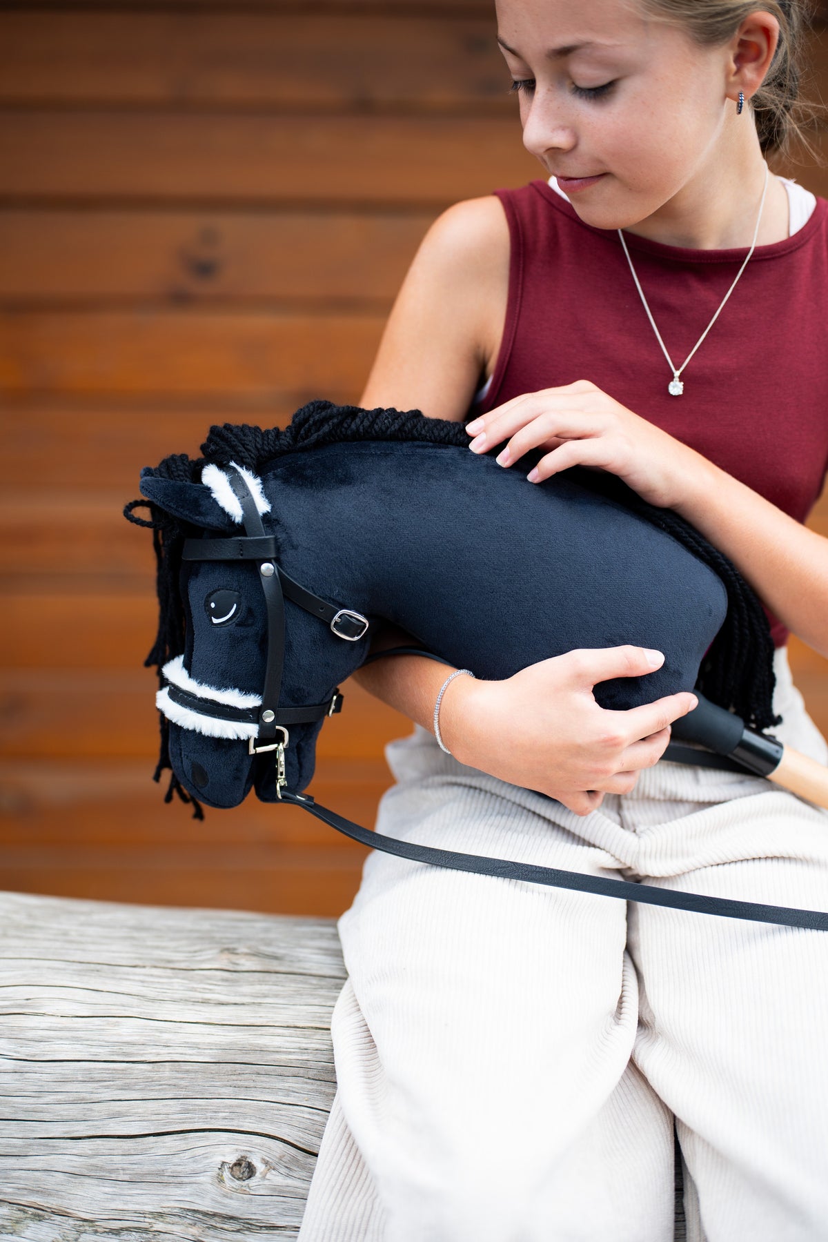 Dívka ve vínovém topu drží Hobby Horse Mahal s černou ohlávkou od LarDen v náručí před dřevěnou stěnou, s černou plyšovou hlavou, černou přízovou hřívou a bílým kožešinovým nánosníkem, s kompletní uzdečkou s kožíškem