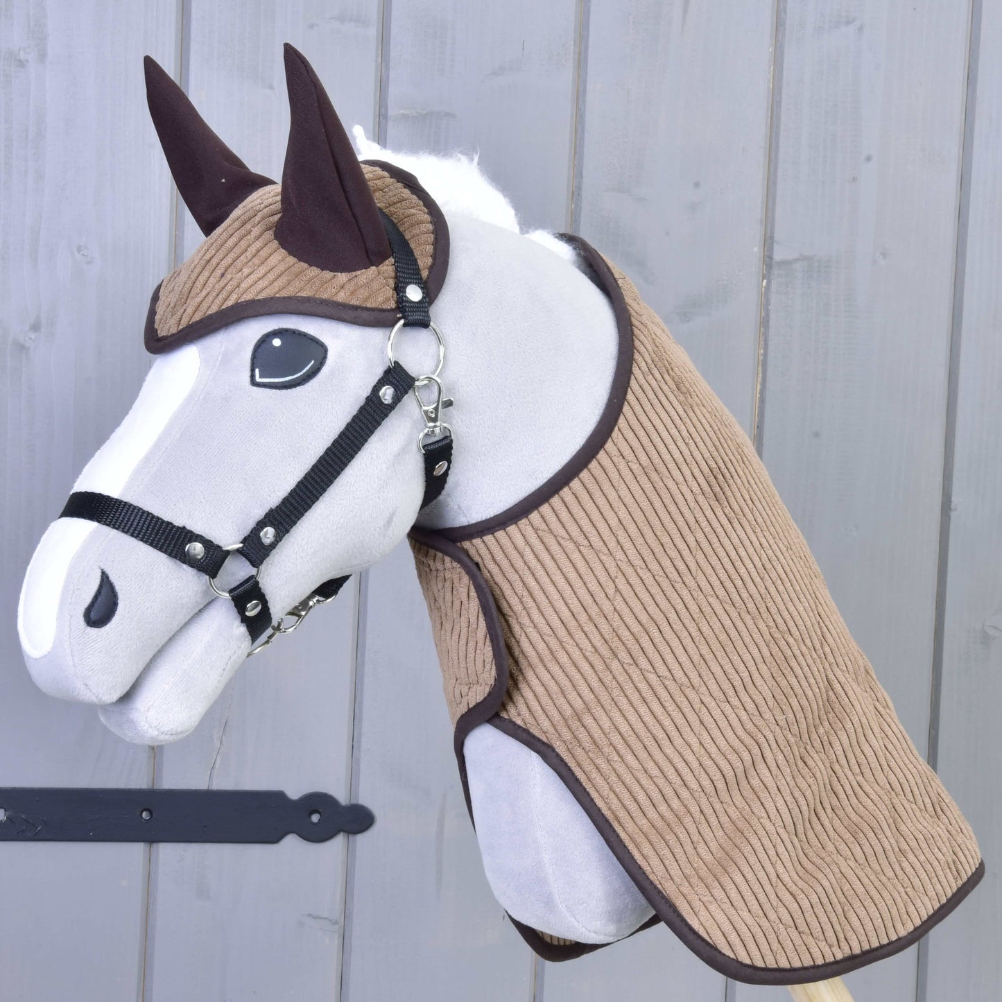 Set Manchester Le Chocolate od Hobby Horse LarDen s plyšovým Hobby Horse s bílým obličejem, černým postrojem, hnědýma ušima a strukturovanou hnědou látkou na hlavě a krku, doplněný Hobby Horse dekou, na dřevěné tyči před šedou dřevěnou stěnou.