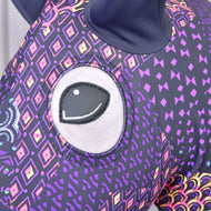 Detail ochranného obleku Hobby Horse ochranný oblek Mandala (Lycra) od Hobby Horse LarDen, zobrazující velké vyšívané oko a fialovou látku s barevnými geometrickými a kosočtvercovými vzory; nahoře je vidět tmavé ucho.