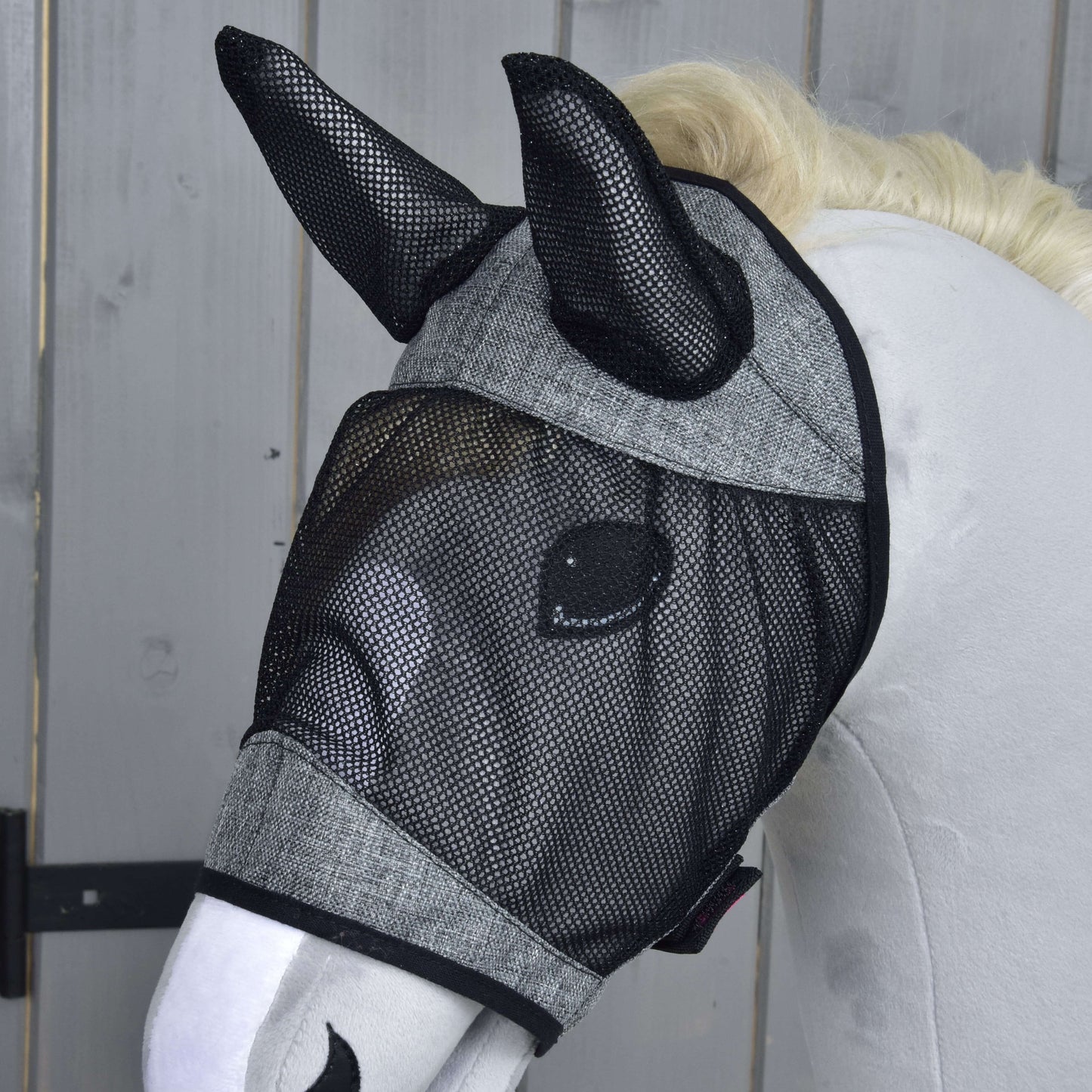 Bílá figurína koně má na sobě masku proti hmyzu de Luxe Light Grey od Hobby Horse LarDen, která zakrývá uši a oči, a stojí před dřevěným pozadím.