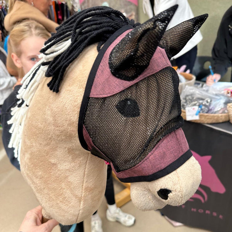Detail ručně vyrobeného Hobby Horse se síťovanou maskou a černo-bílou přízovou hřívou. V rozmazaném pozadí lidé a stánek s koňskými předměty ukazují kreativního ducha Hobby Horsing.