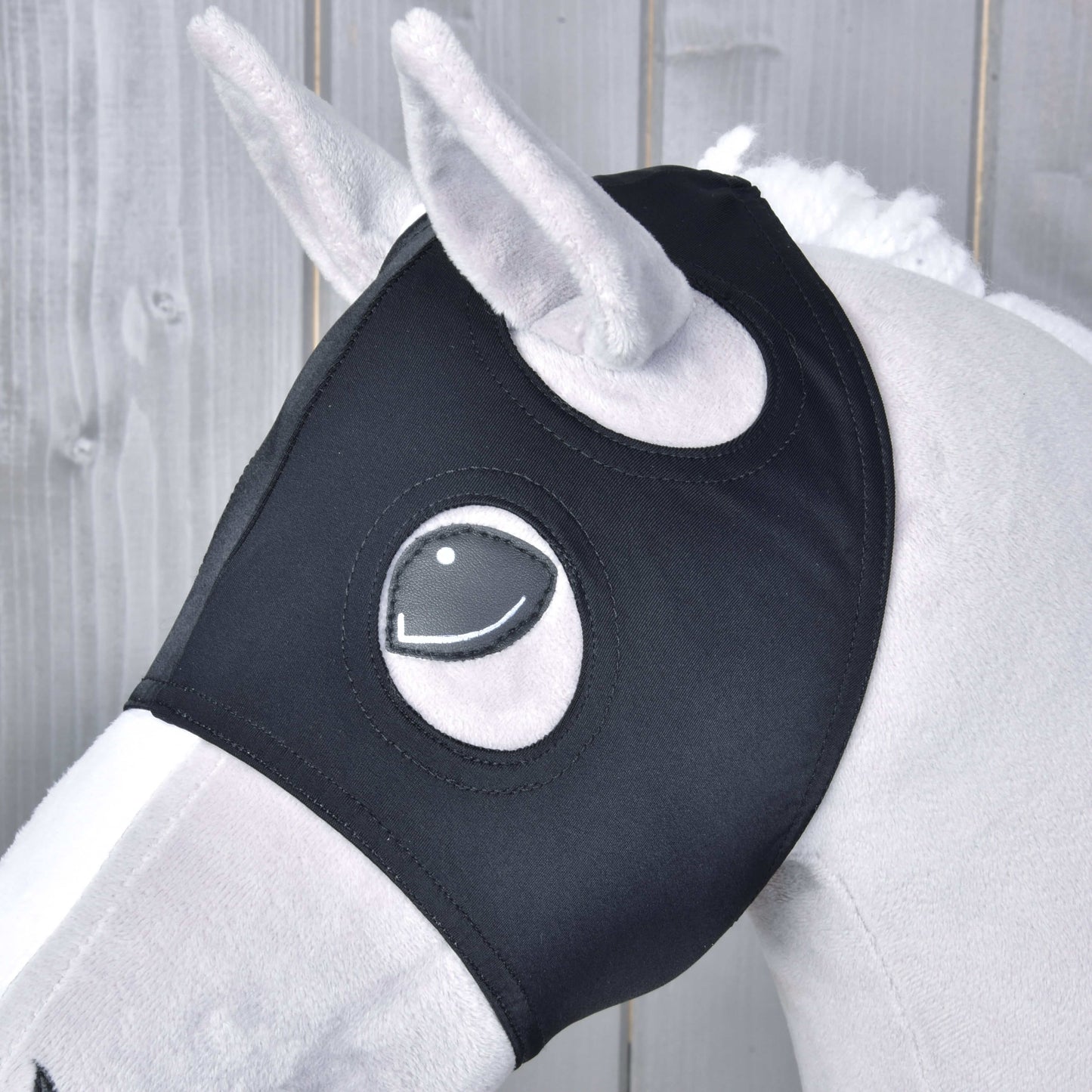 Detail bílého plyšového koně s maskou Hobby Horse LarDen „Horse Mask without ears Flamingo“, z materiálu PES s otvory pro oči. V pozadí jsou dřevěné panely.