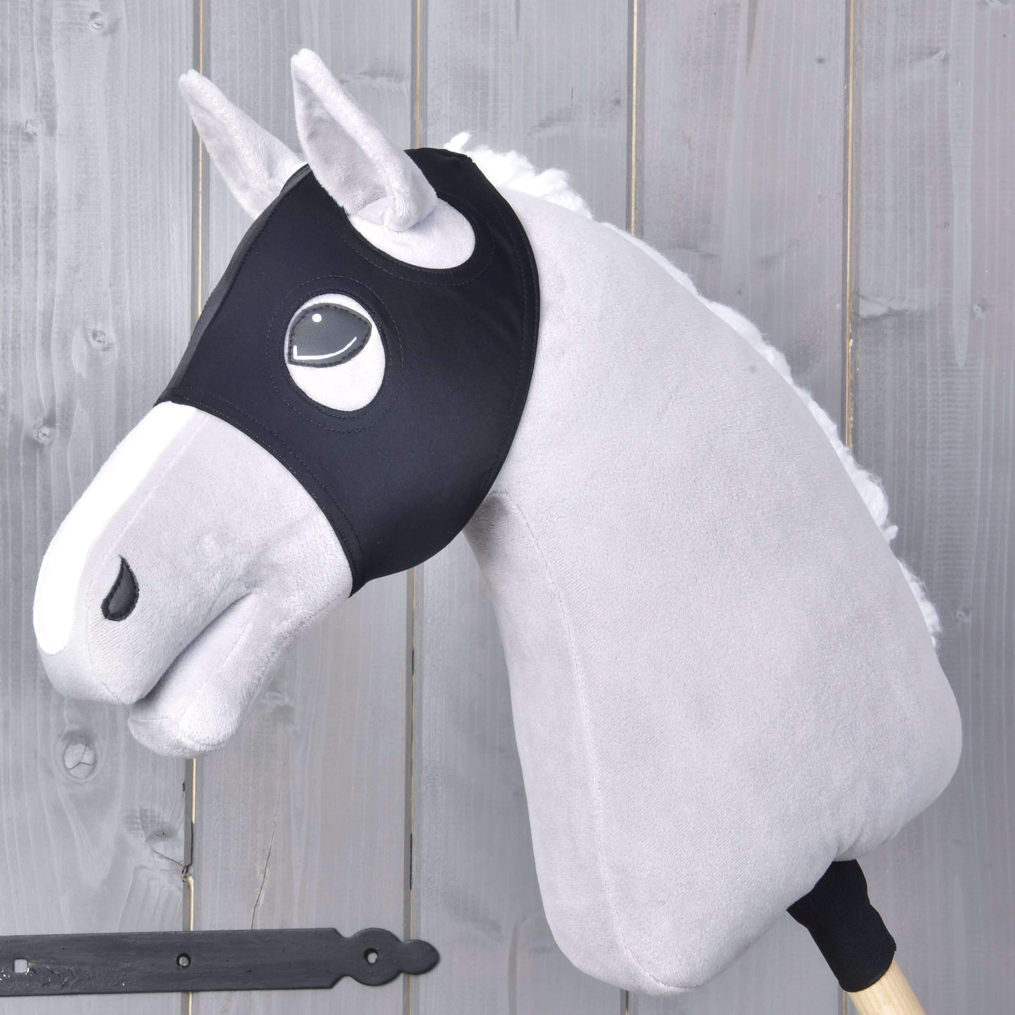 Plyšový Hobby Horse se světle šedou srstí, bílou hřívou a černou uzdečkou má na sobě masku Maska bez uší Flamingo od Hobby Horse LarDen a je nasazený na dřevěné tyči před pozadím z šedých dřevěných panelů.