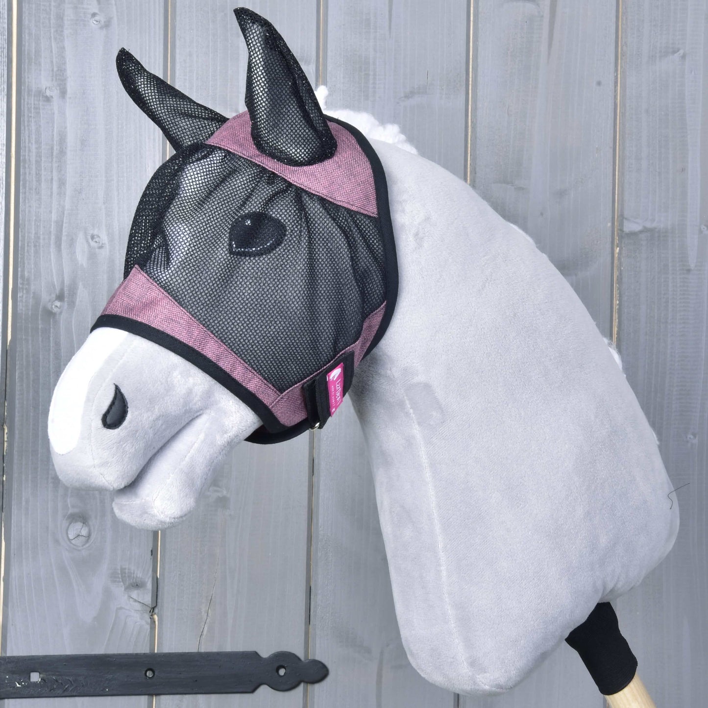 Maska proti hmyzu de Luxe Pink od Hobby Horse LarDen na šedém plyšovém Hobby Horse s růžovým lemováním a pratelnou maskou proti hmyzu, nasazeném na dřevěné tyči, na pozadí z dřevěných prken.